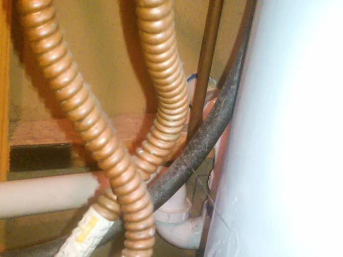 Corrugated copper-colored water heater connectors and a white tank in a utility closet